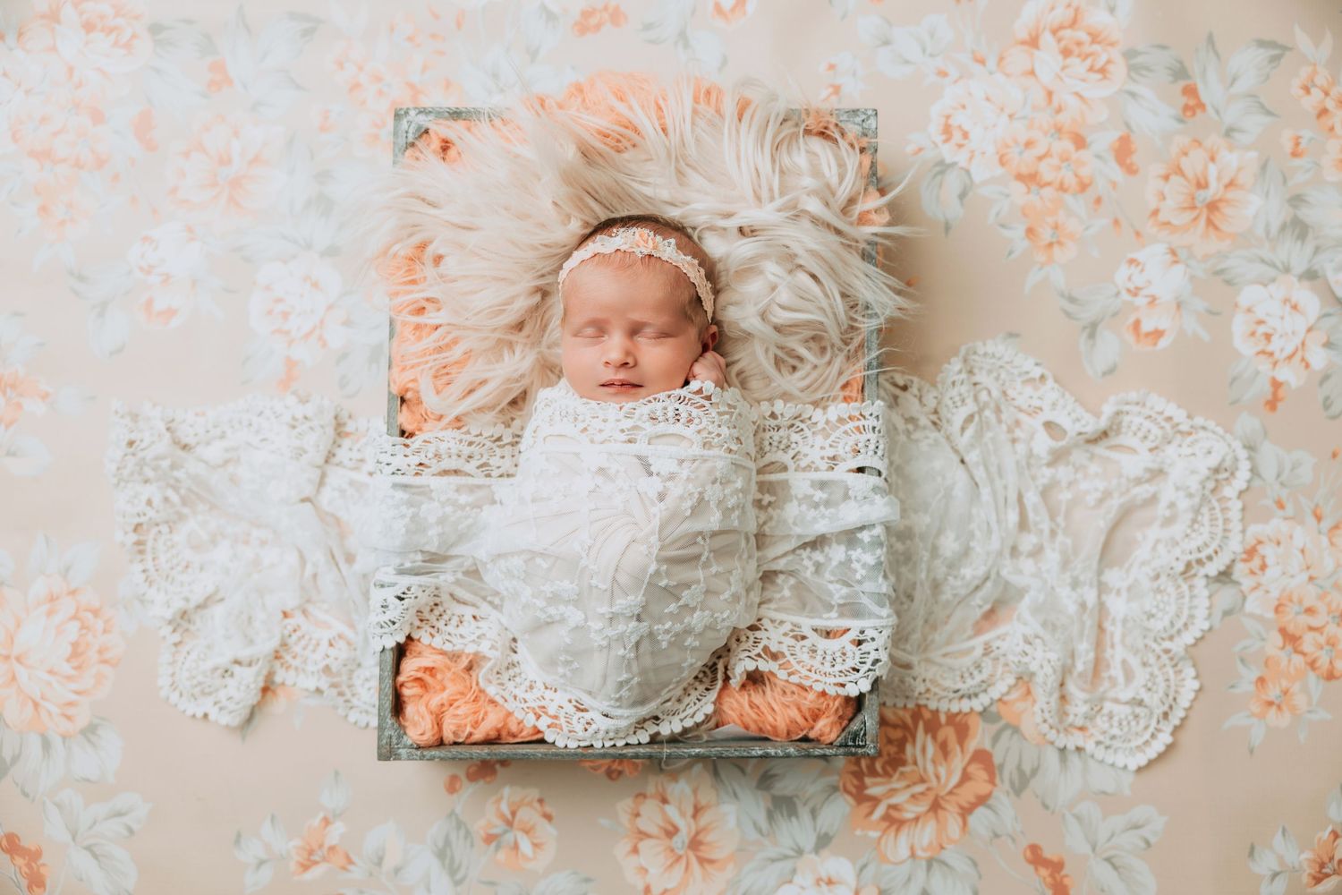 Newborn baby swaddled in white lace wrap, sleeping in a rustic wooden box with peach floral backdrop.