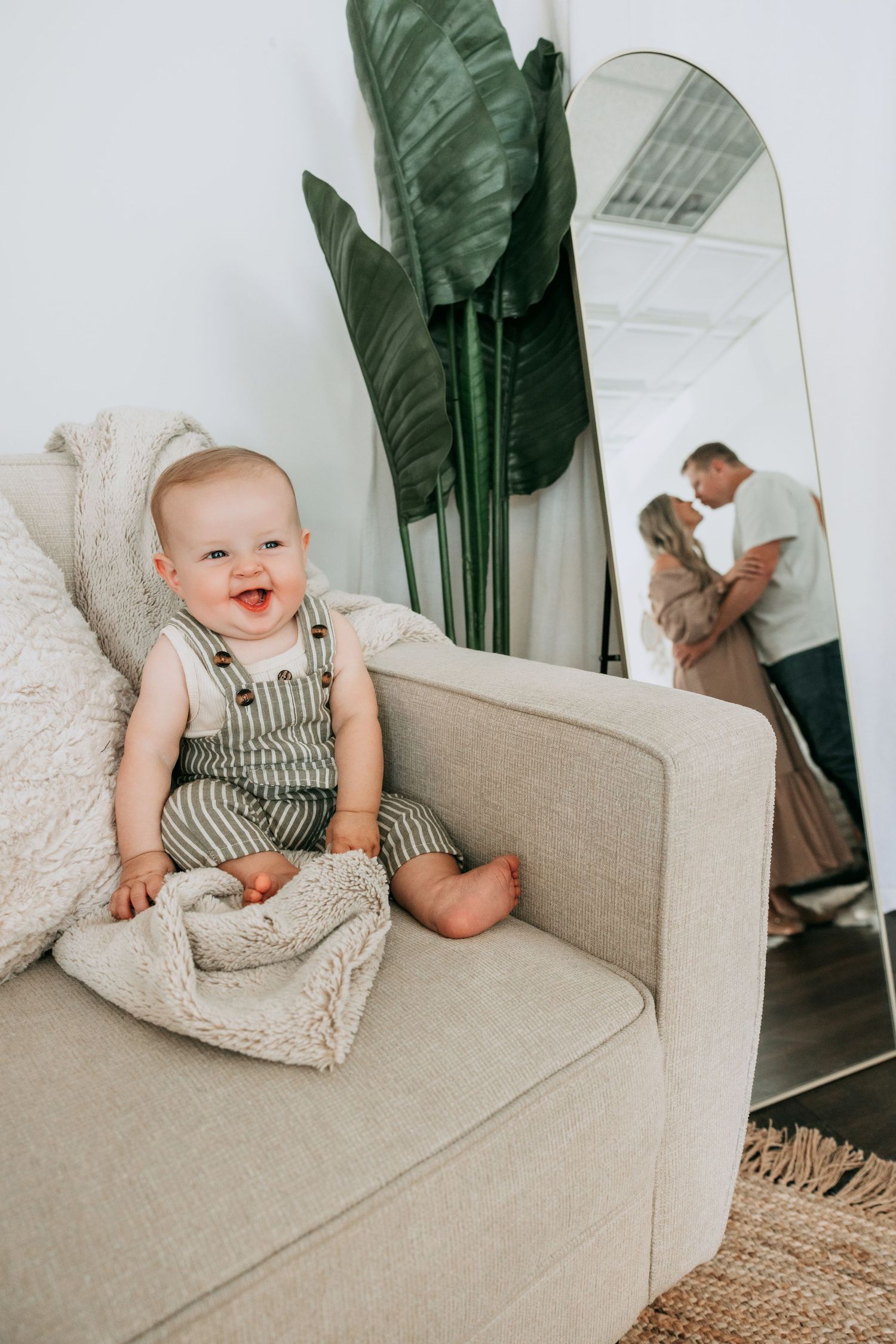 A smiling baby in striped overalls sits on a beige armchair, with parents embracing in a mirror reflection behind them.