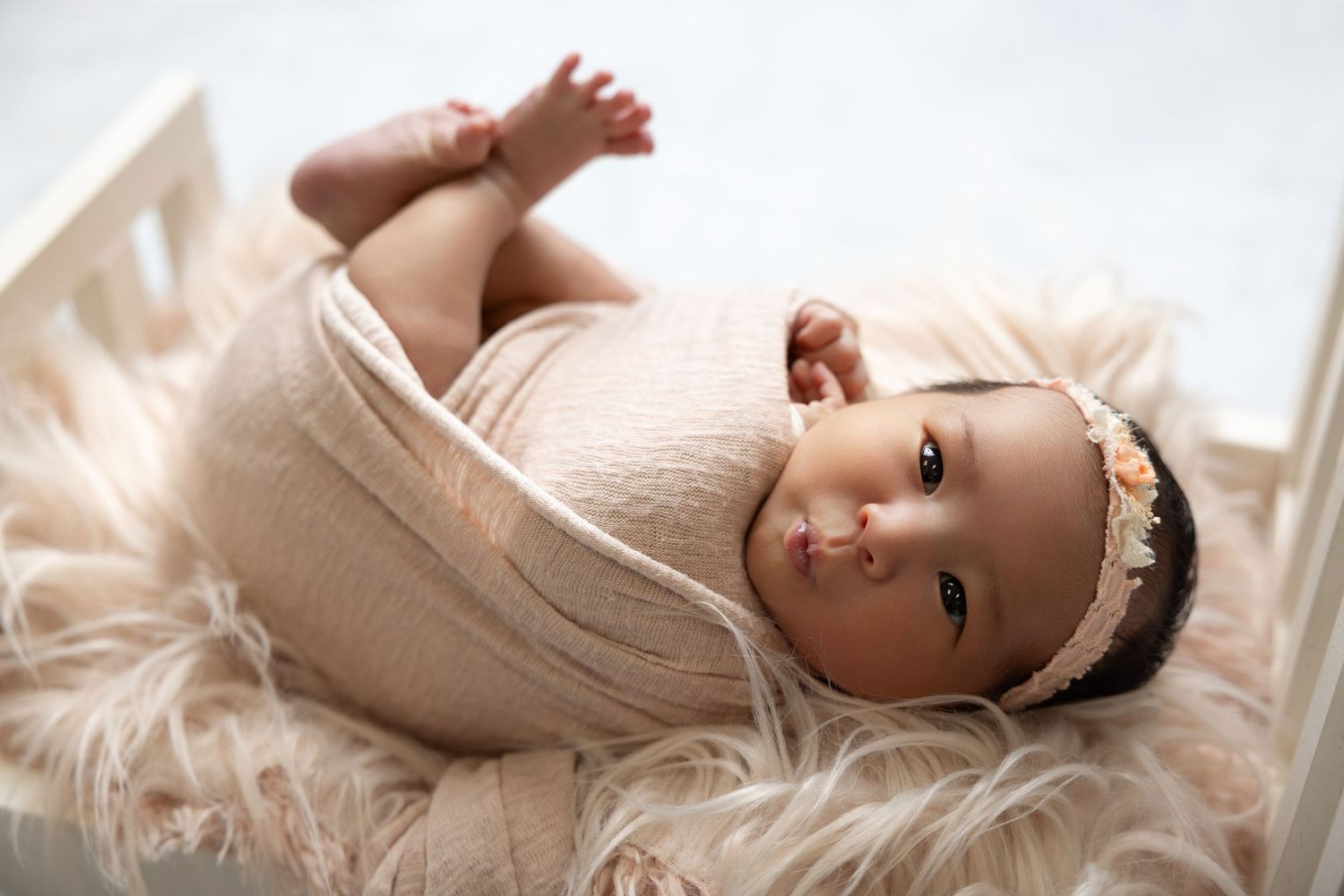 Newborn baby girl wrapped in blush pink knit swaddle with floral headband lying in a white wooden crib.