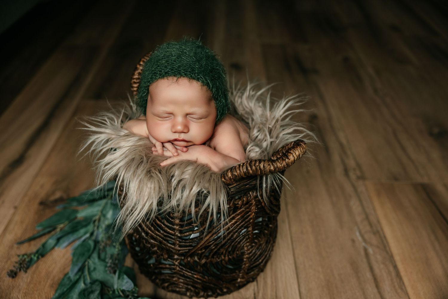 Sleeping newborn baby in green knit bonnet nestled in wicker basket with fluffy fur, on wood floor.