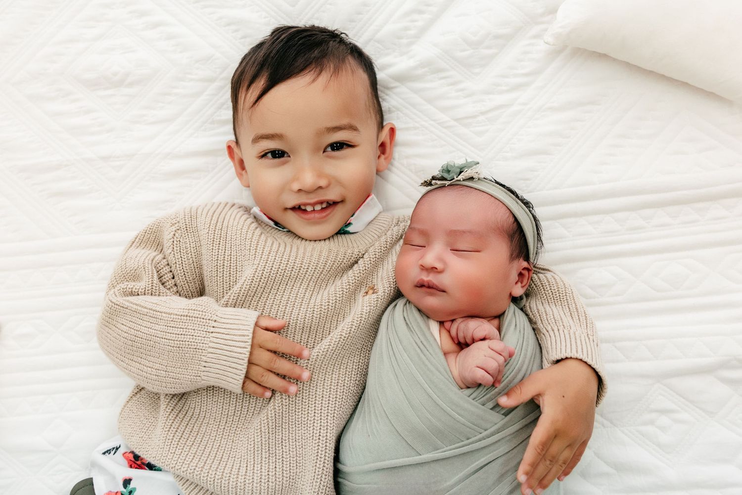 Smiling toddler in beige sweater poses with sleeping swaddled newborn baby on white bedding.