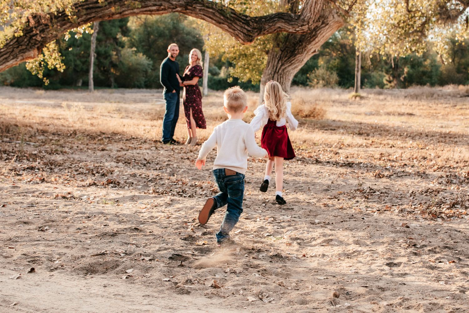 Two young children running toward parents standing under large tree in dry autumn field at the Bonsall Preserve.