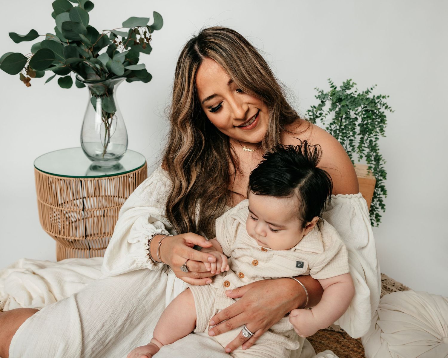 Smiling young mother in white dress cuddling dark-haired baby in bright boho studio setting.
