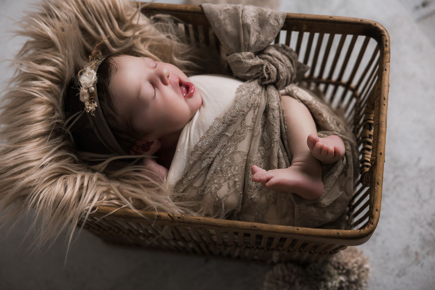 Newborn baby girl sleeping in wicker basket wrapped in lace with floral headband on fur blanket. Fallbrook Newborn Photographer