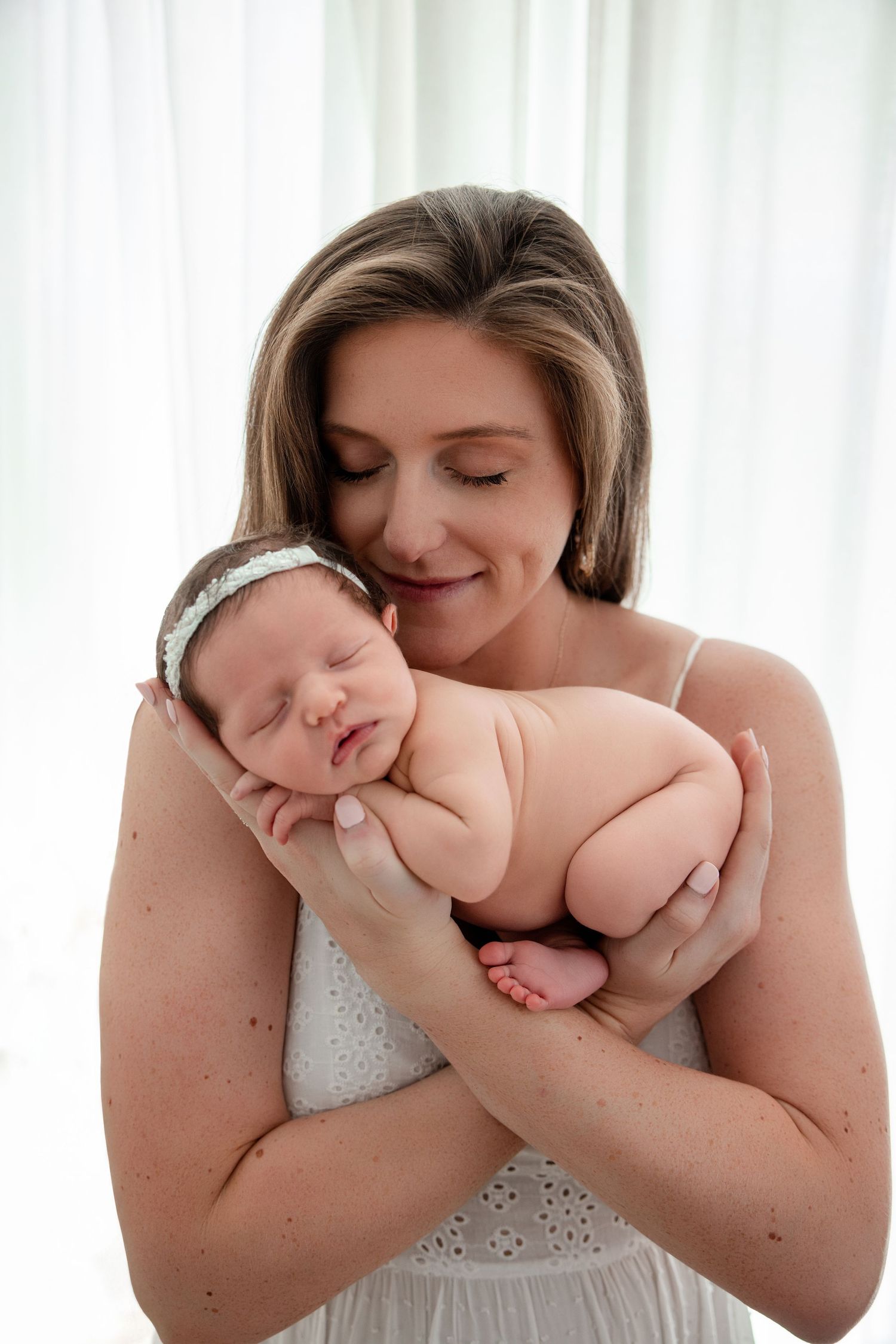 A smiling mother in a white dress tenderly holds her sleeping newborn baby against a soft white background.