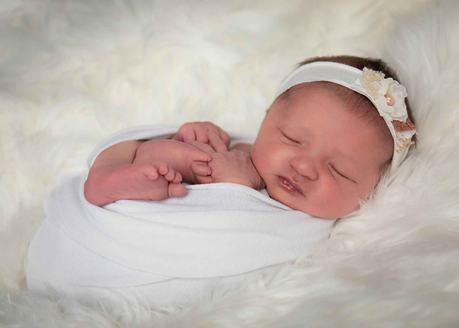 Peaceful newborn baby sleeping wrapped in white swaddle with floral headband on fluffy white fur blanket.