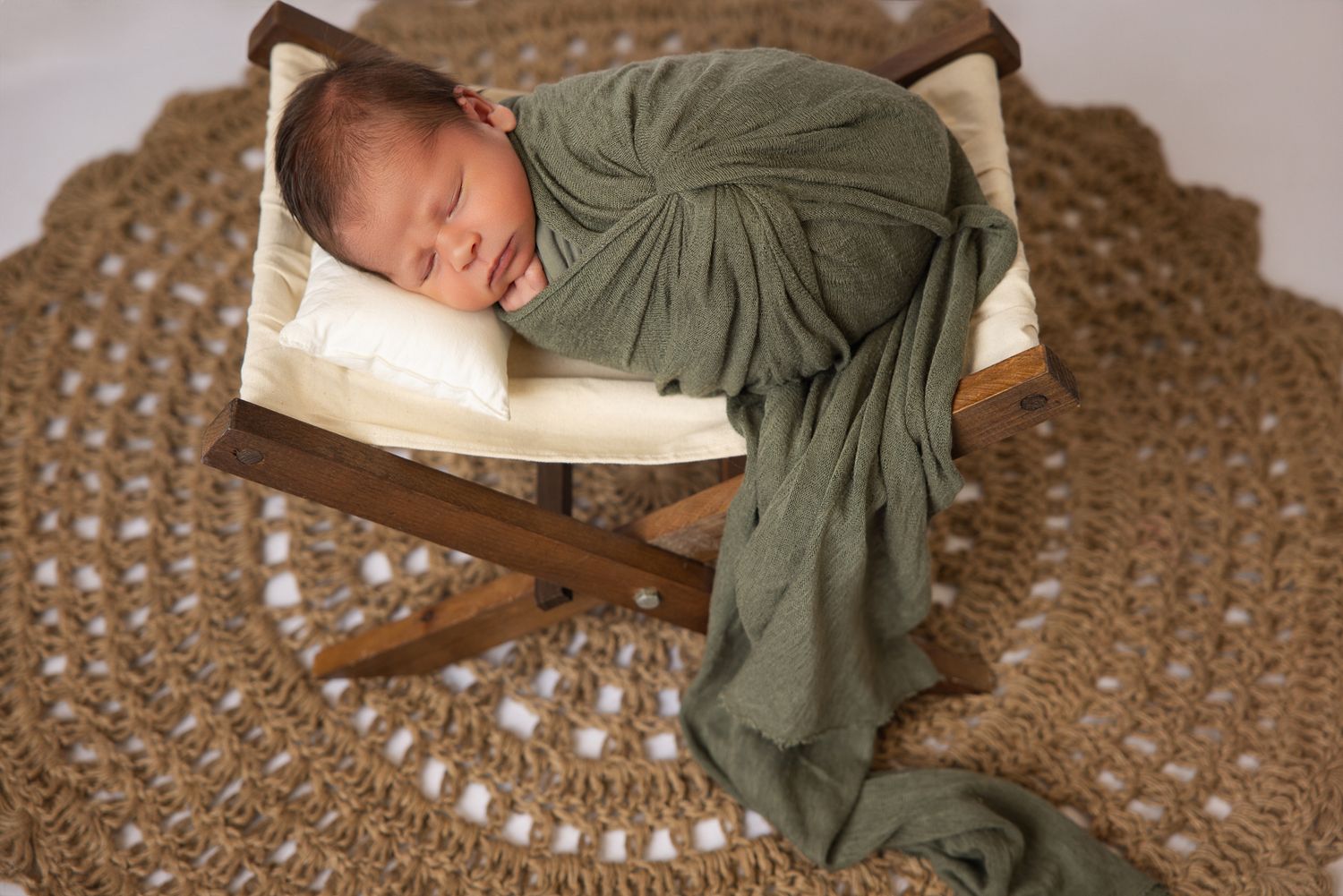 Newborn baby swaddled in green wrap sleeping peacefully on a tiny wooden bed atop a round woven rug.