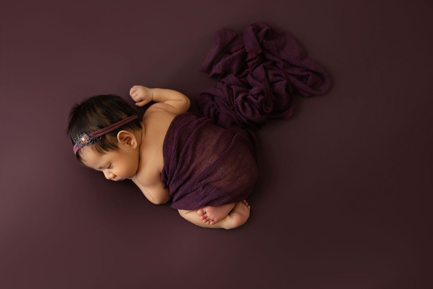 Newborn baby girl sleeping peacefully on a dark plum background, wrapped in a matching purple fabric.