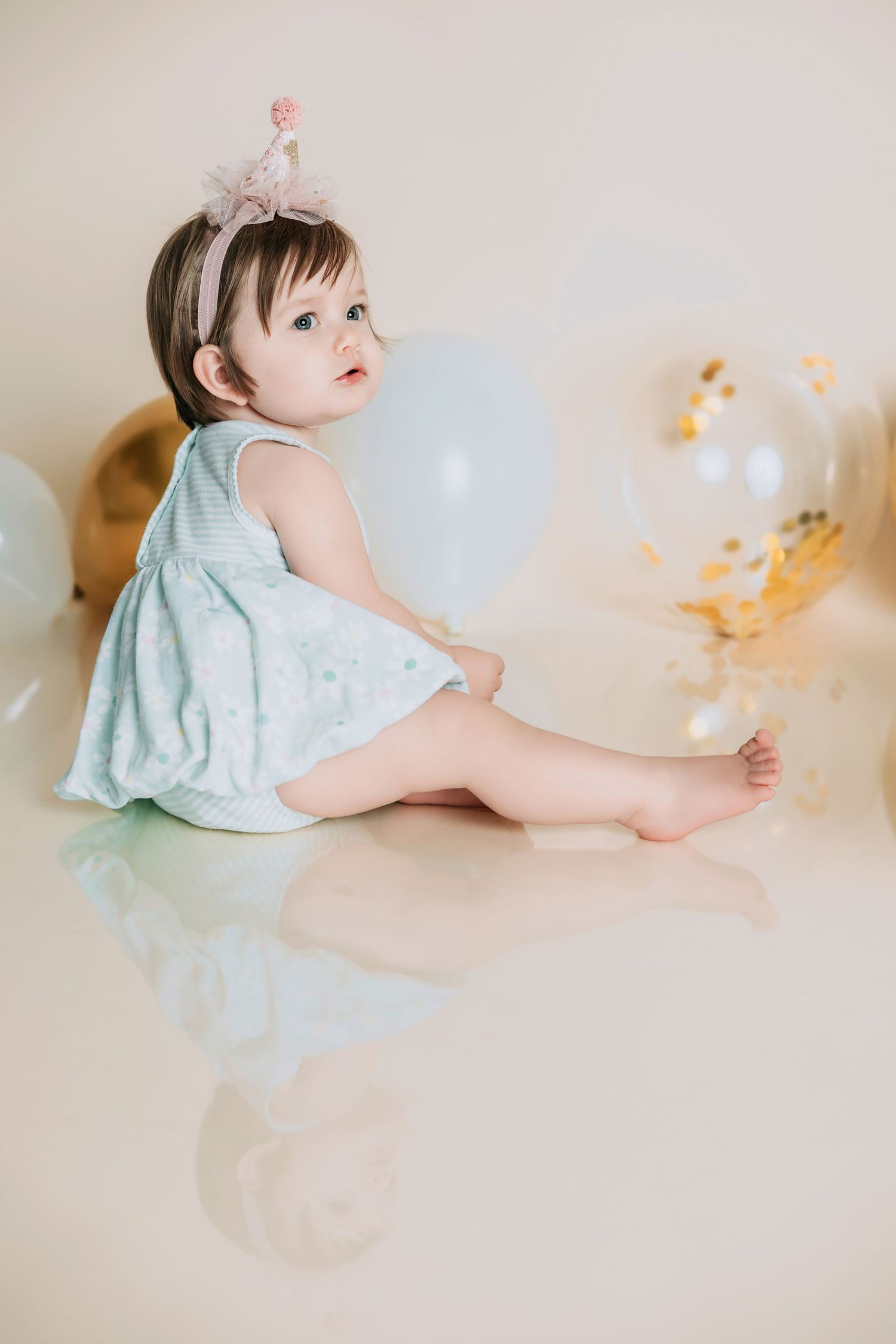 A baby girl in a mint dress and pink crown headband sits on a reflective white floor surrounded by pastel balloons.
