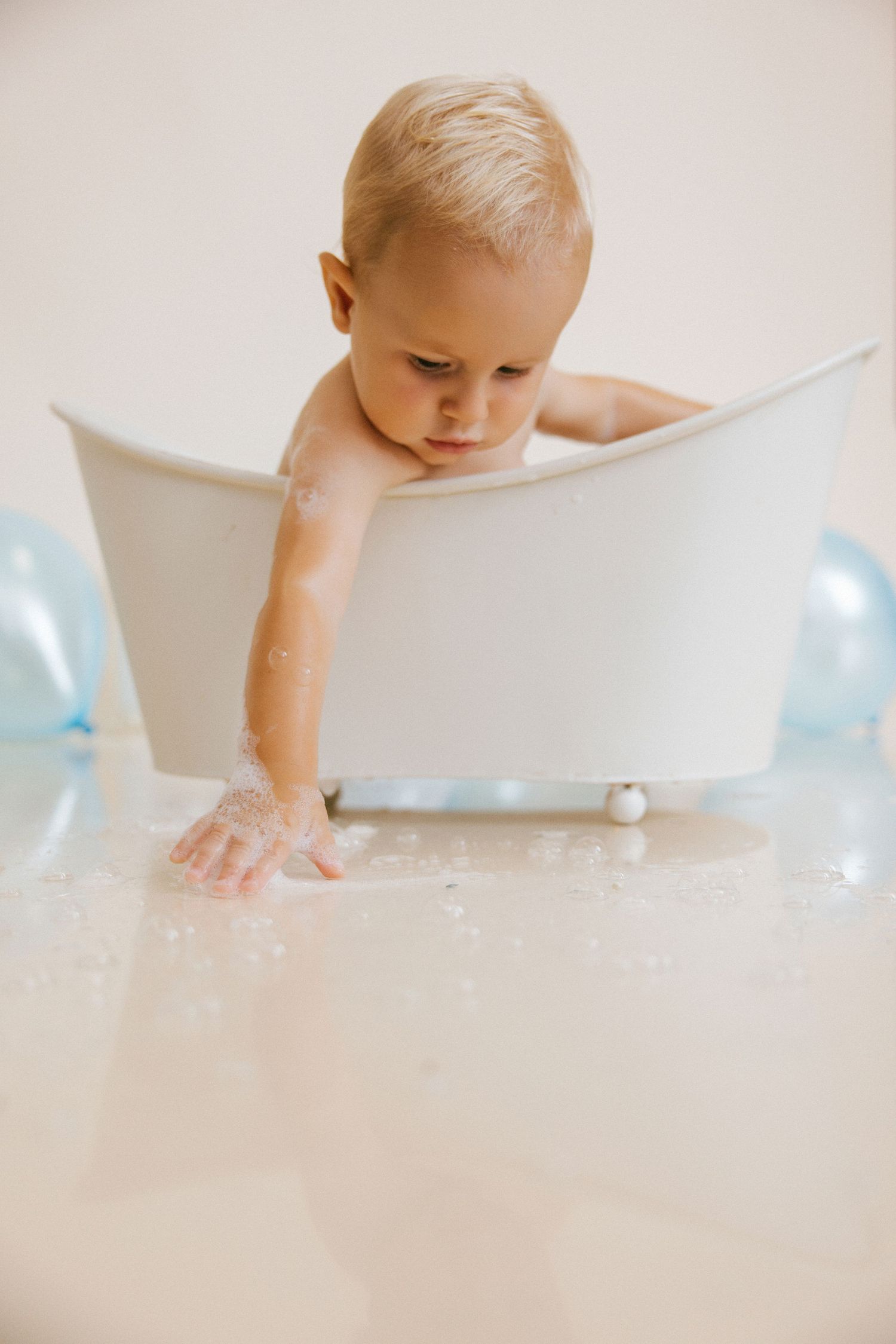 A cute blonde toddler playing in a small white bathtub with soap suds and blue balloons nearby.