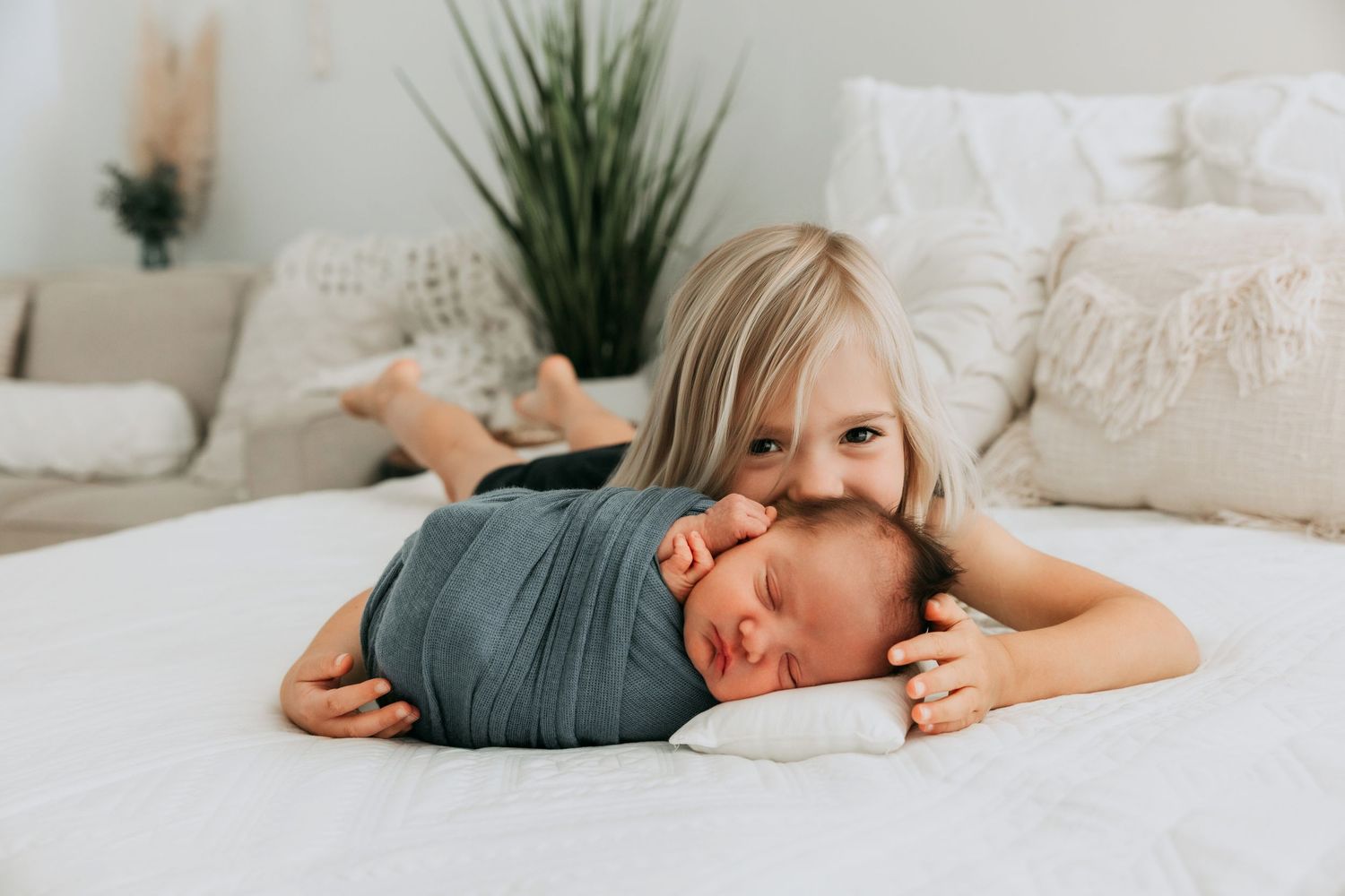 Young blonde girl lying on white bed cuddling a swaddled newborn baby sibling.