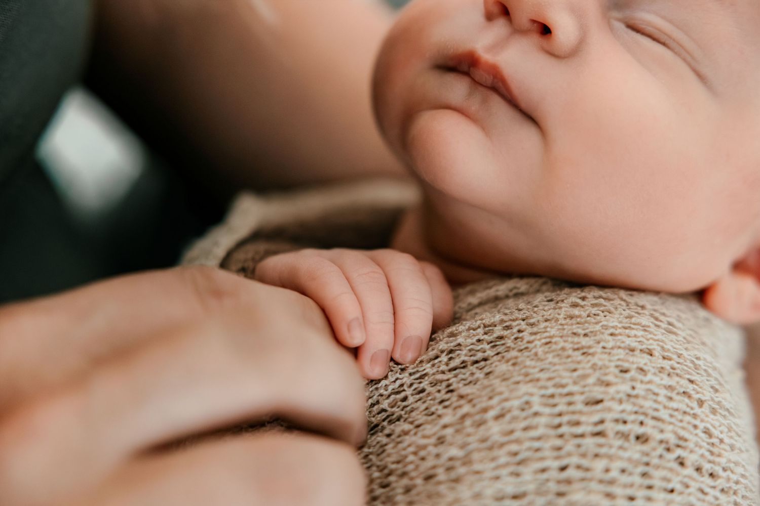 Peaceful sleeping newborn baby wrapped in beige knit blanket, tiny hand resting on parent's finger.