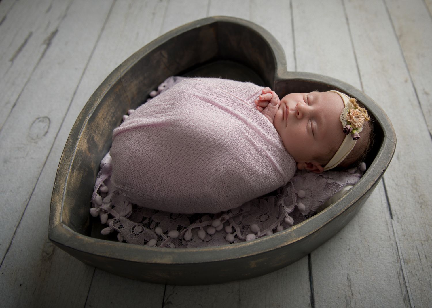 Newborn baby swaddled in pink wrap, sleeping in a wooden heart-shaped bowl with a floral headband.