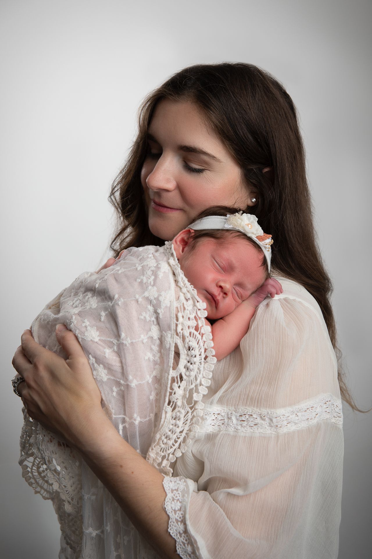 A young mother in white lace gown tenderly holds her sleeping newborn baby wrapped in soft white fabric.