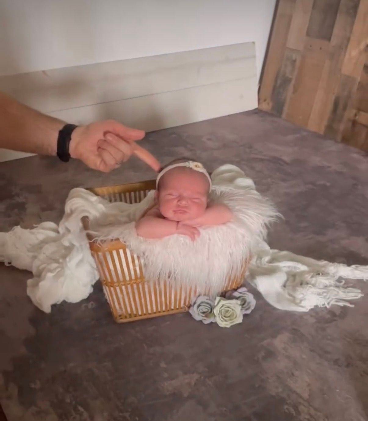 Newborn baby sleeping in a wicker basket with white fur, wearing a floral headband, during a professional photo shoot.