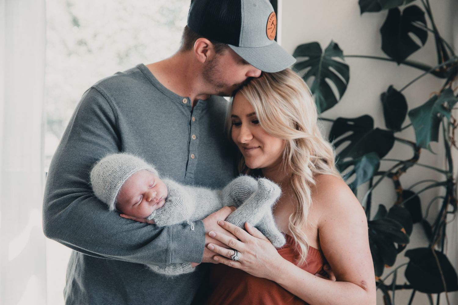 A couple tenderly holds their newborn baby wrapped in a gray knit blanket near a window with tropical plants.