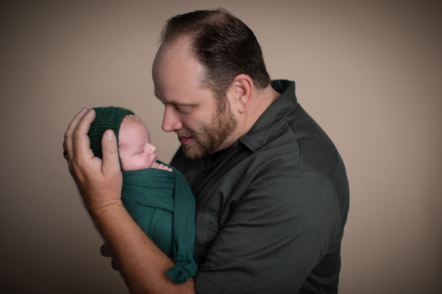 A bearded father tenderly holds his newborn baby wrapped in a green swaddle against a neutral background.