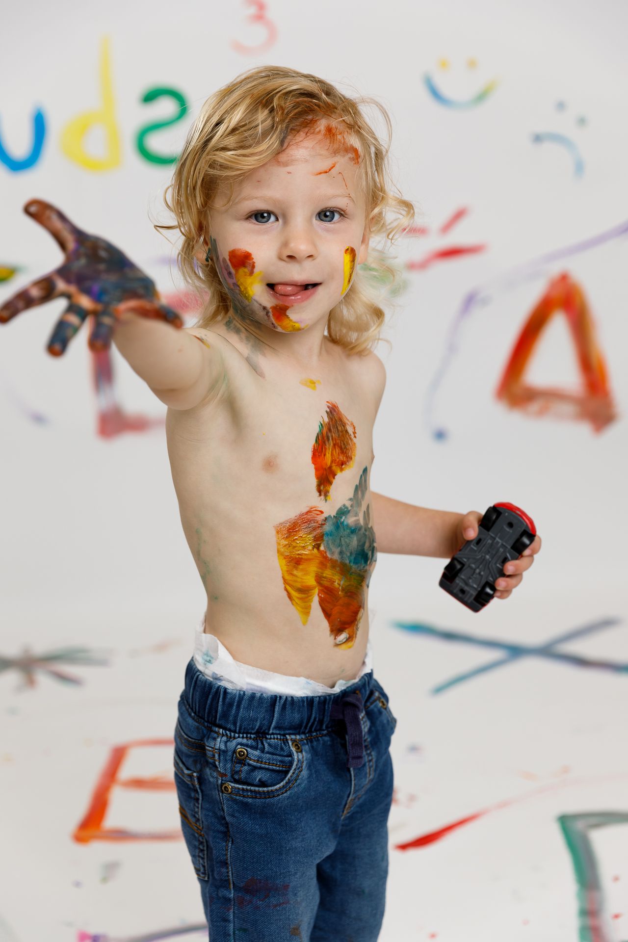 Toddler boy covered in colorful paint holding toy car, standing in front of painted wall.