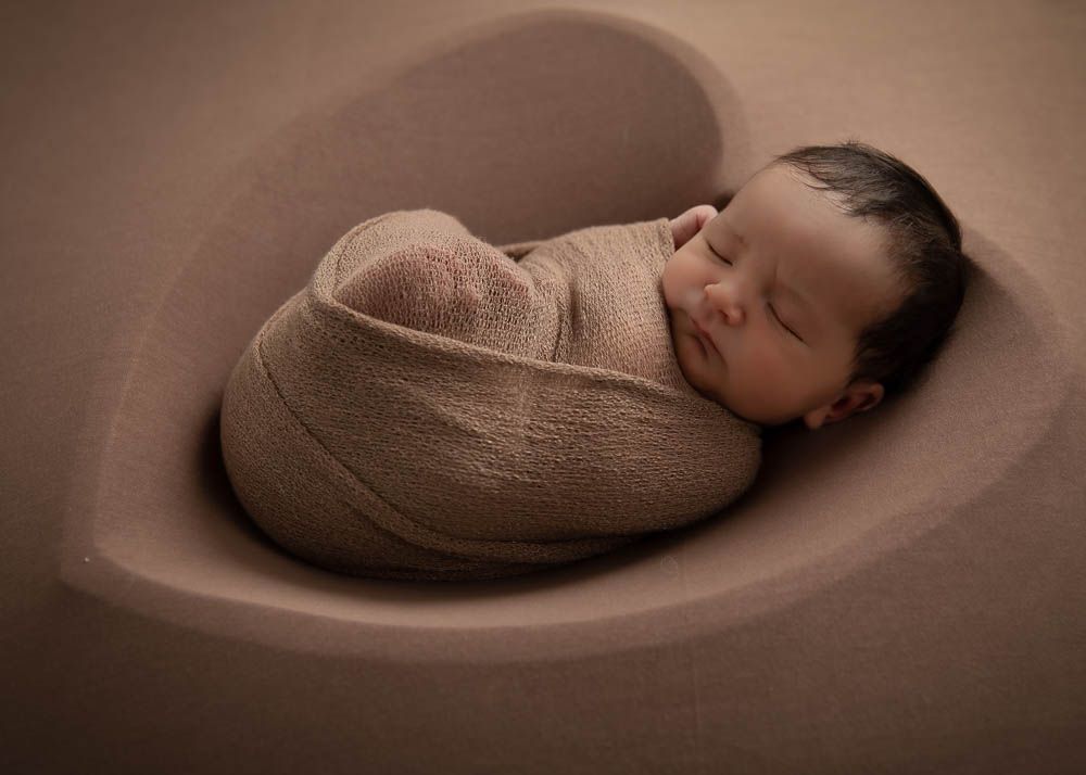 Sleeping newborn baby swaddled in beige wrap, nestled in a heart-shaped bowl for a studio portrait.