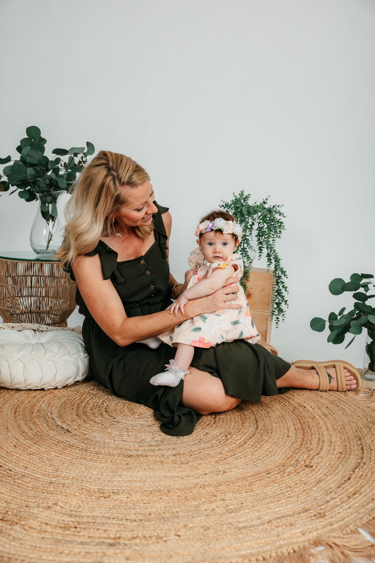 A blonde mother in a black dress sits on a jute rug holding her baby girl in a floral outfit surrounded by greenery in the Bonsall Studio