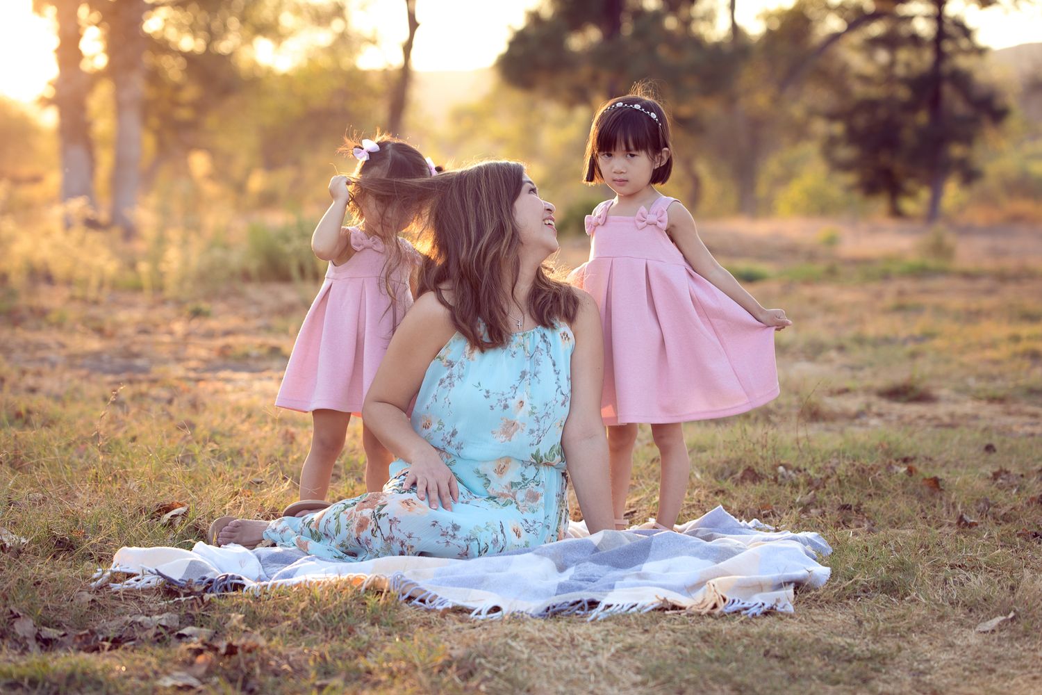 A mother sits on a blanket outdoors as her two young daughters in pink dresses stand beside her playing with her hair in Bonsall, CA