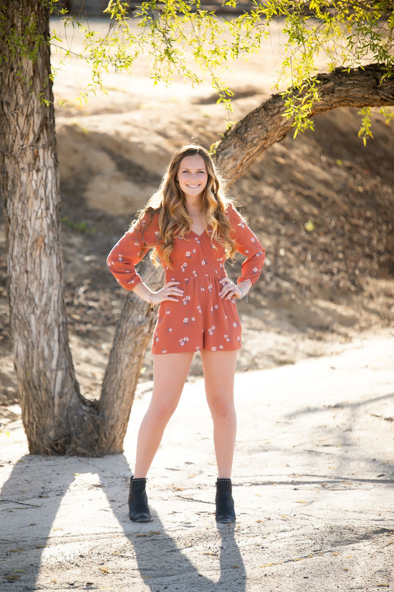 Smiling woman in rust floral romper poses with hands on hips under tree in sunlight.