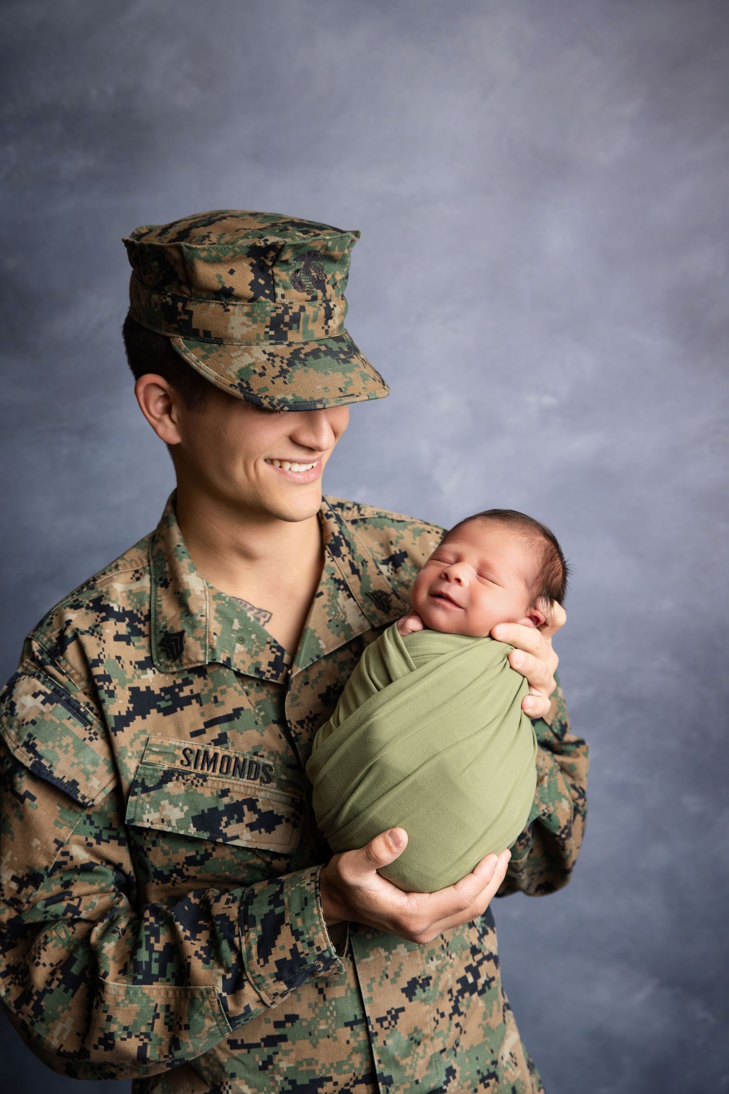 Smiling female Marine in camouflage uniform holding swaddled newborn baby during military portrait session.