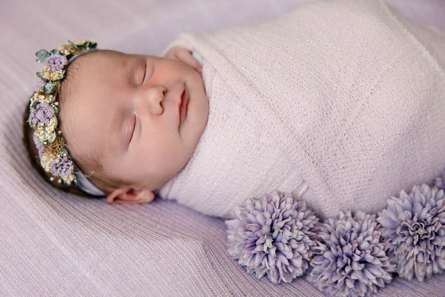 Peaceful newborn baby girl swaddled in lavender wrap with floral crown, lying on purple blanket.