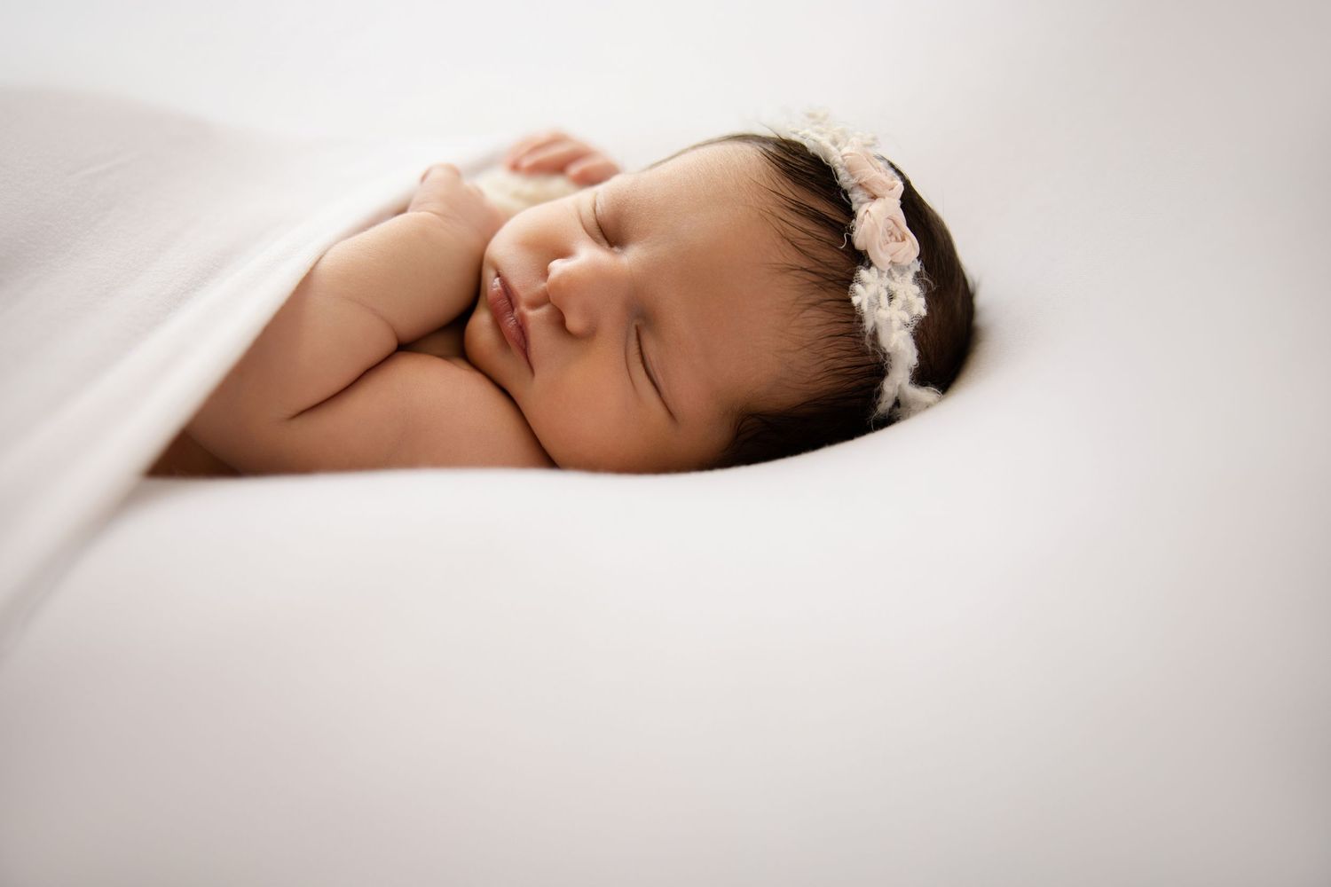 Peaceful newborn baby girl sleeping on white blanket wearing a delicate floral headband in a professional photo.