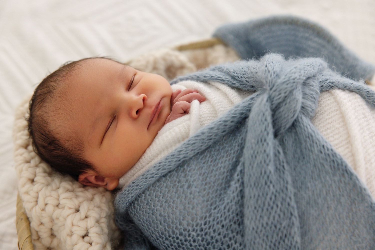 Sleeping newborn baby swaddled in a soft blue knit blanket, resting peacefully in a cozy basket.