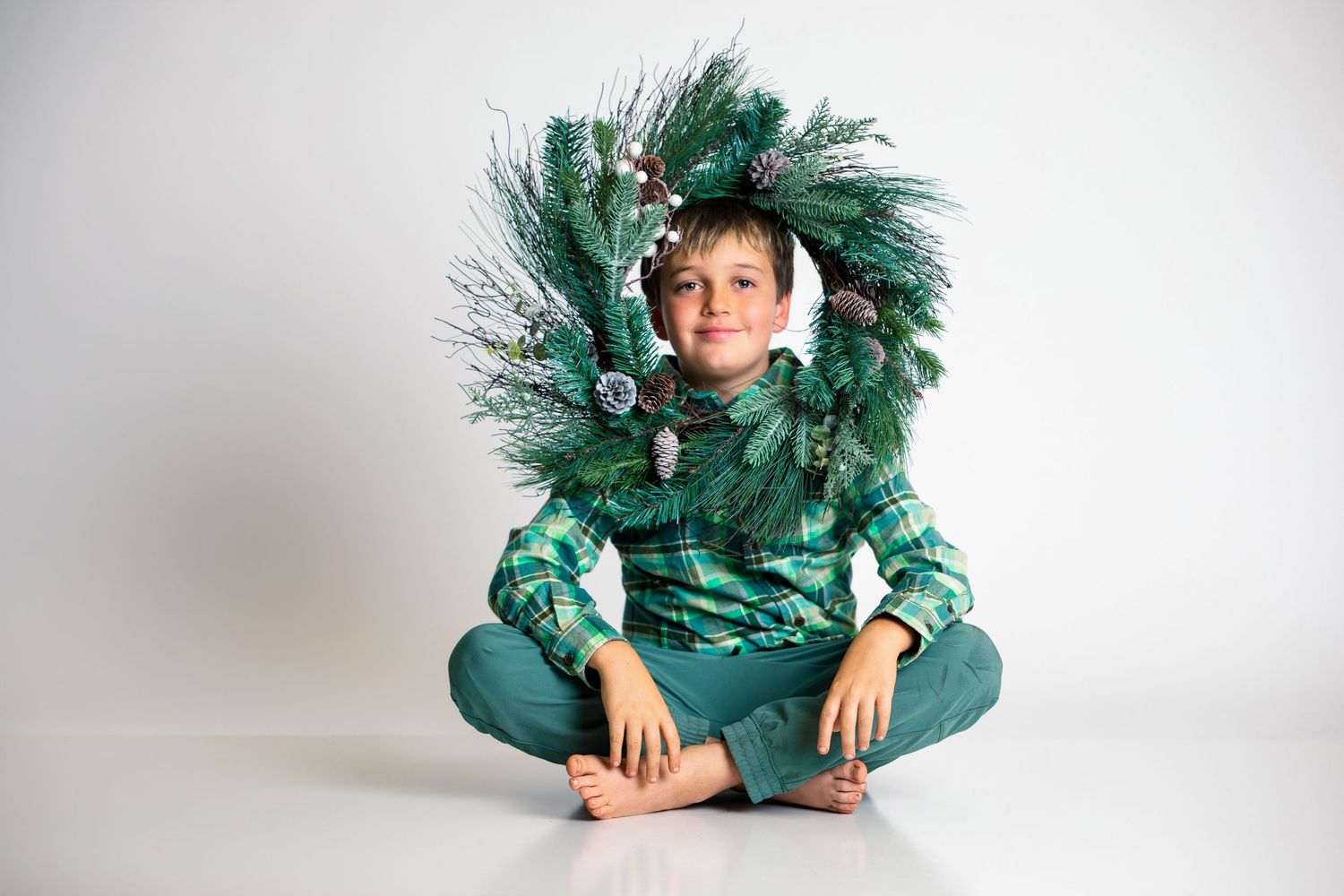 Boy in green plaid shirt sits cross-legged wearing a festive Christmas wreath as a costume on white background.