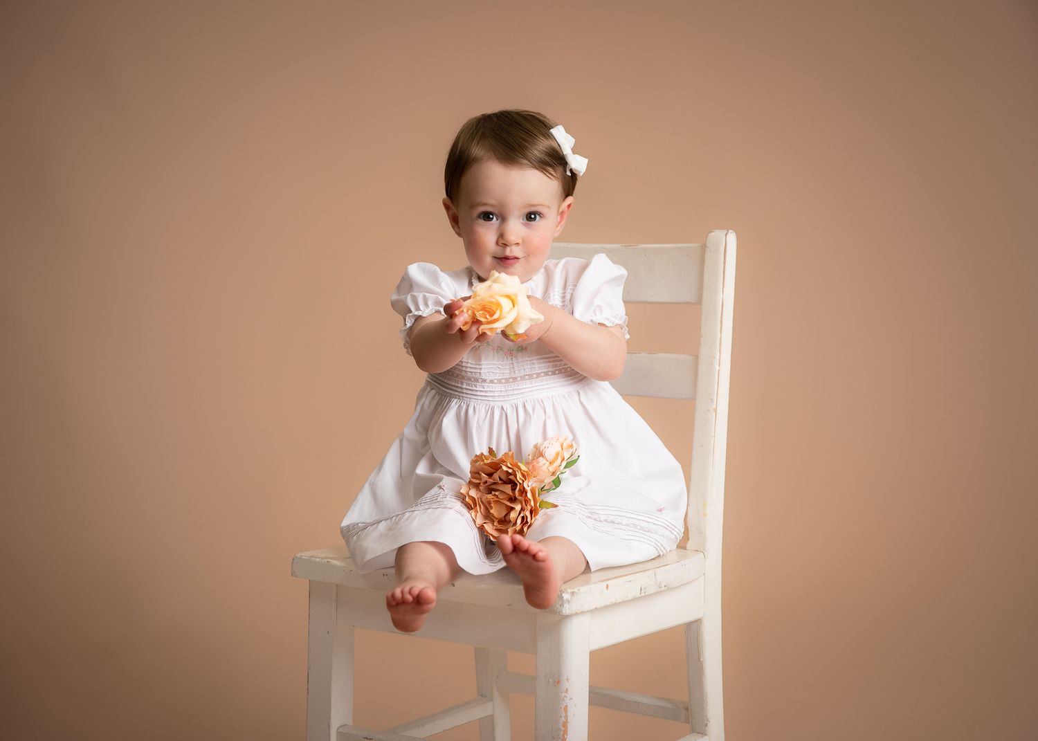 Sweet toddler girl in white dress holding orange rose bouquet on white chair.