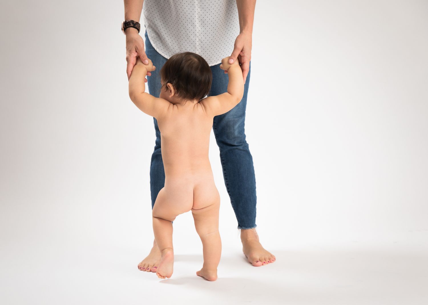 Baby taking first steps while adult holds hands from behind in studio.