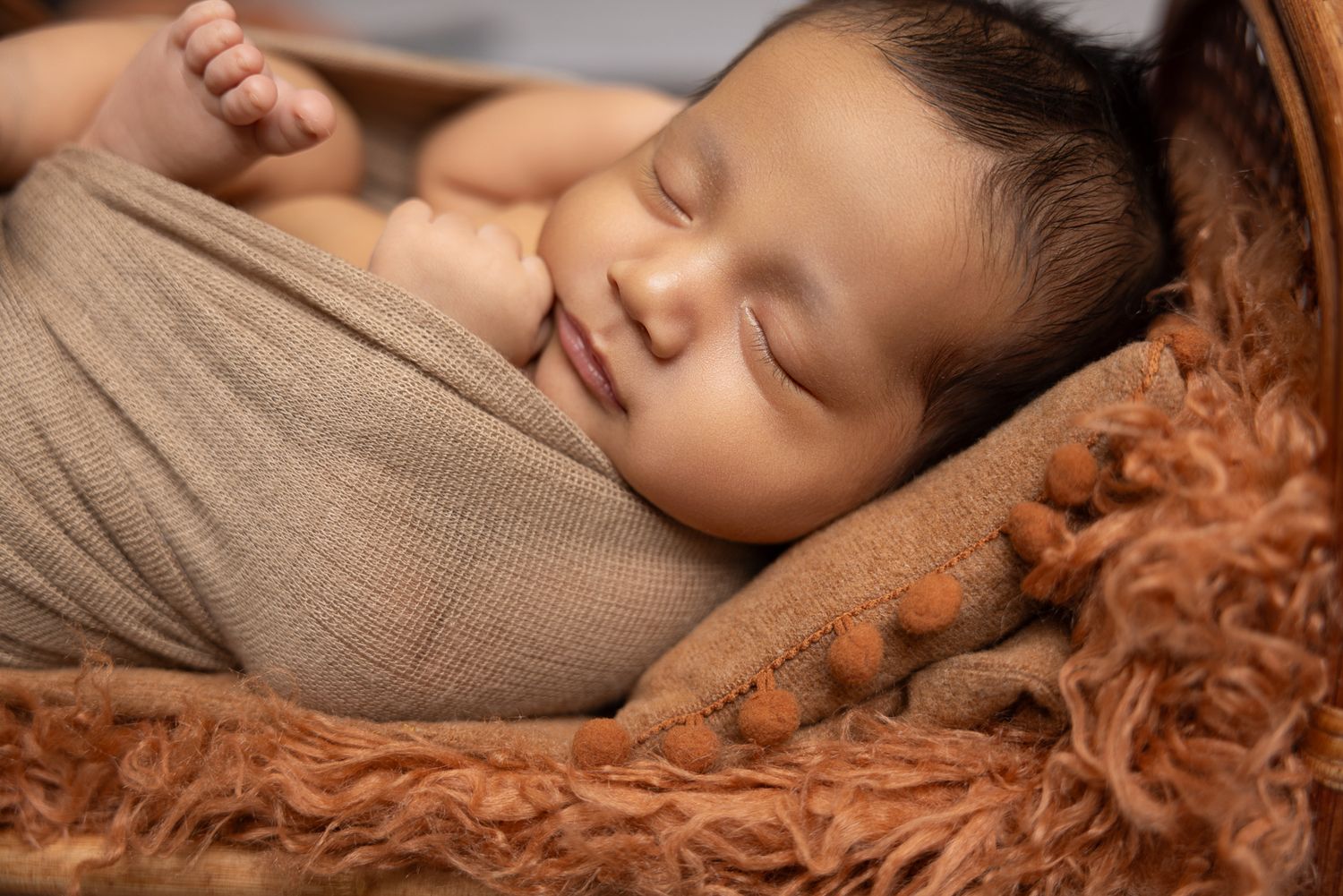 Smiling sleeping newborn baby swaddled in tan wrap resting on warm brown fluffy fur in basket at The Studio Bonsall, near Fallbrook