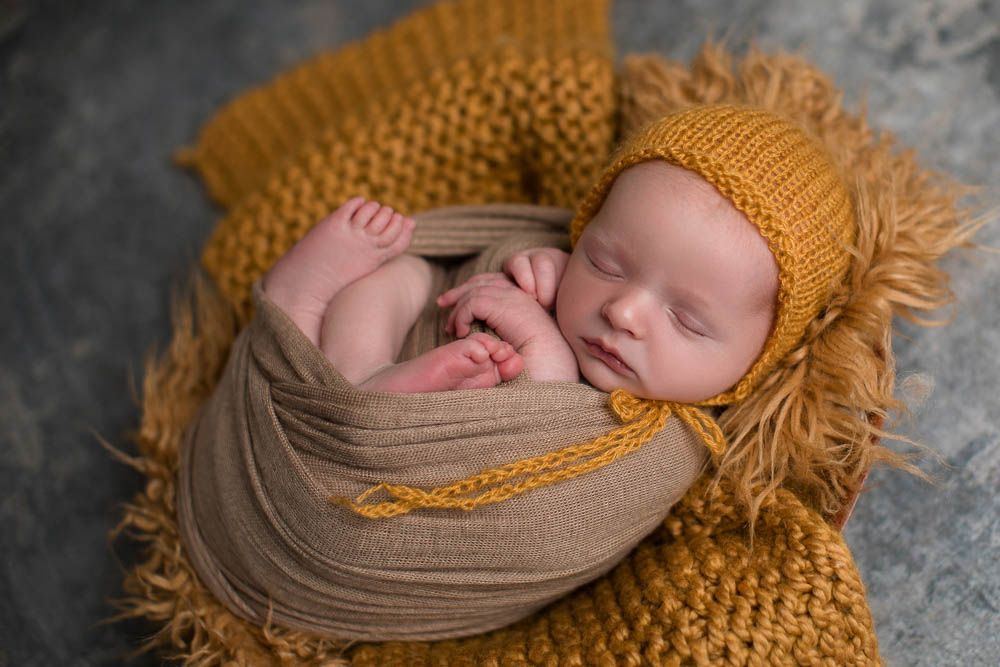 Newborn baby swaddled in mustard yellow wrap and knit bonnet, sleeping in a cozy posed portrait in the Bonsall Studio near Fallbrook, Vista