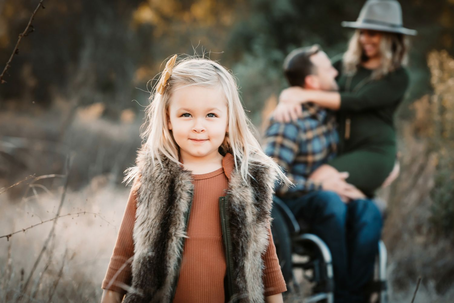 A smiling blonde girl in a fur vest poses in a field with her parents in the background.