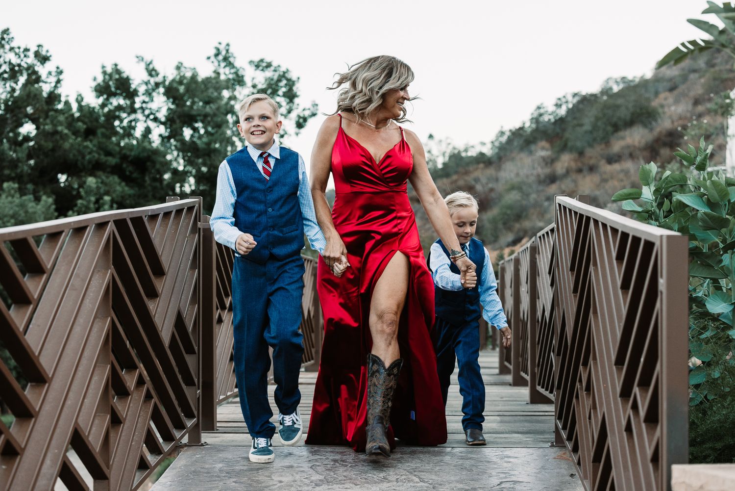 Woman in red dress walks with two boys in suits on a wooden bridge outdoors at Pala Mesa Resort in Fallbrook