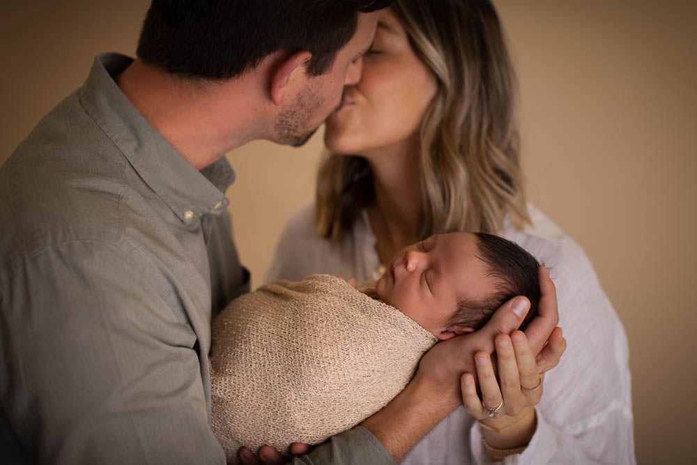 Romantic couple in warm lighting share intimate moments in a series of tender embraces and kisses while holding their newborn