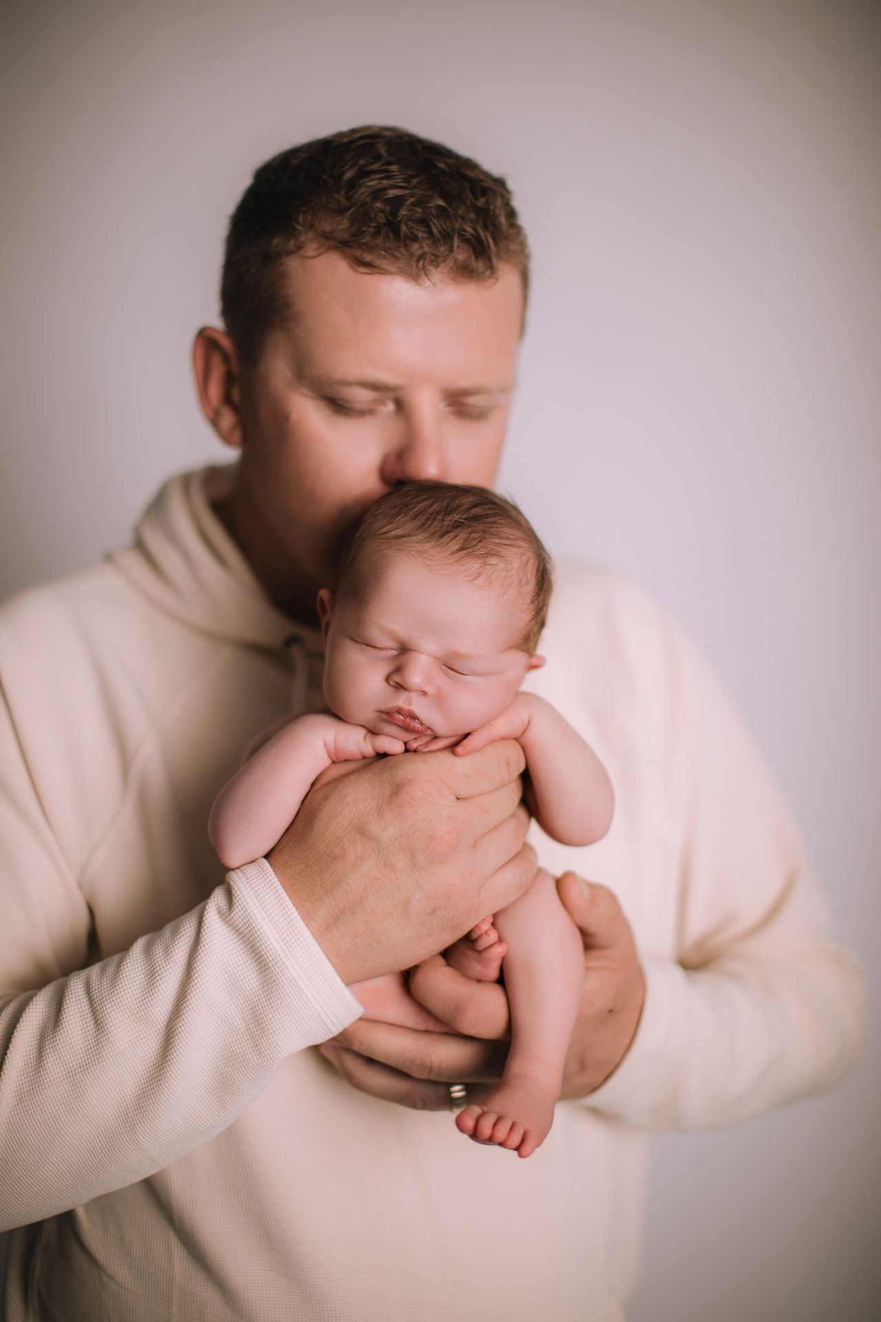 A father in a cream sweatshirt tenderly kisses his sleeping newborn baby cradled gently in his hands.