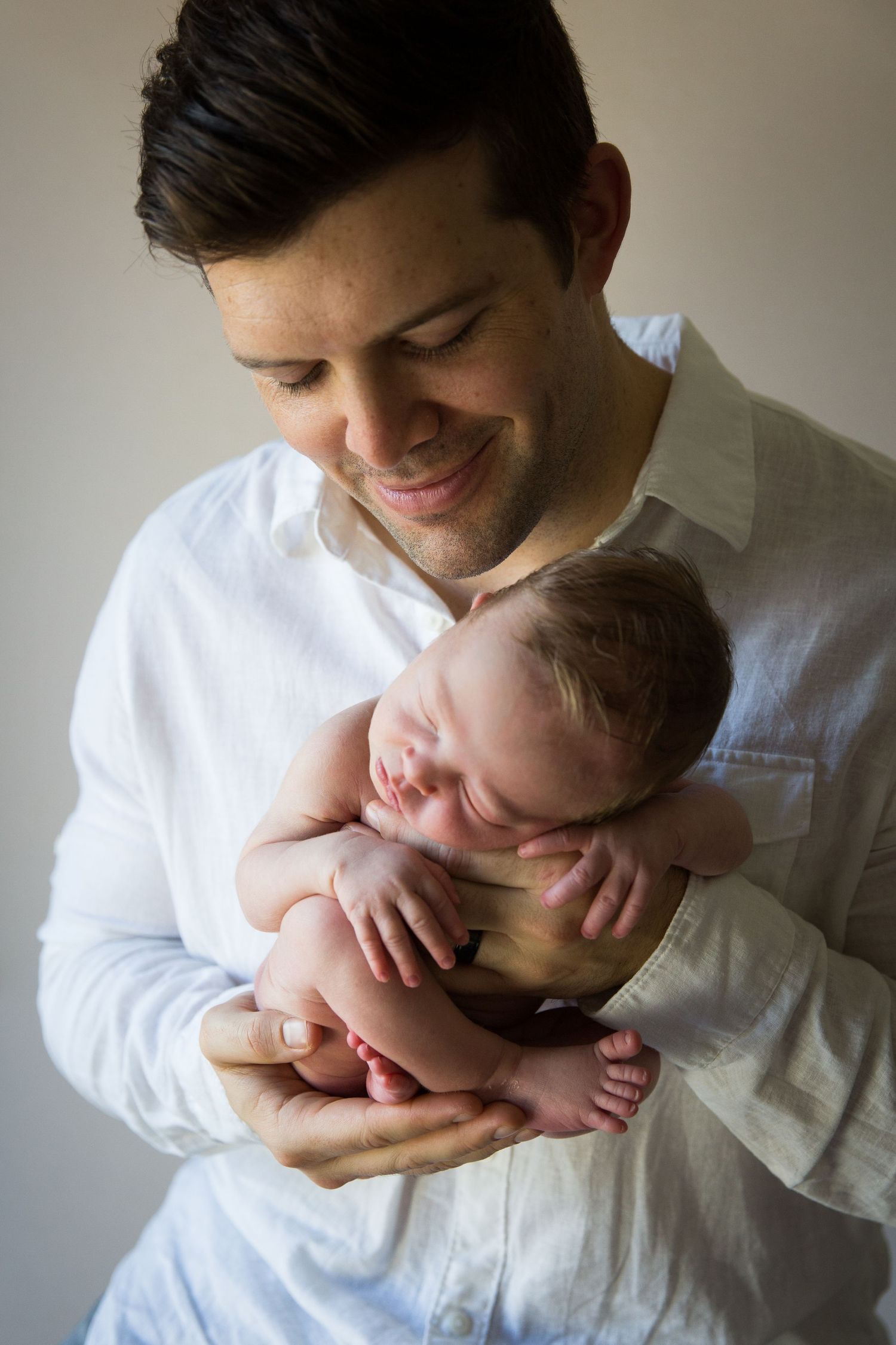 Smiling father in white shirt tenderly holds sleeping newborn baby against his chest.