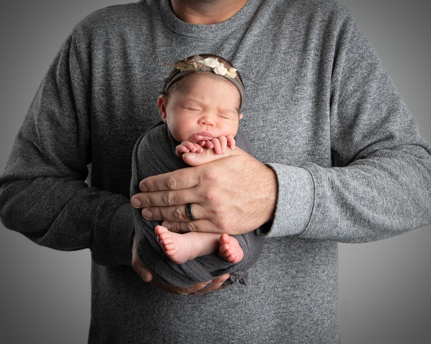 A newborn baby swaddled in gray cloth, cradled gently in an adult's hands, wearing a floral headband.