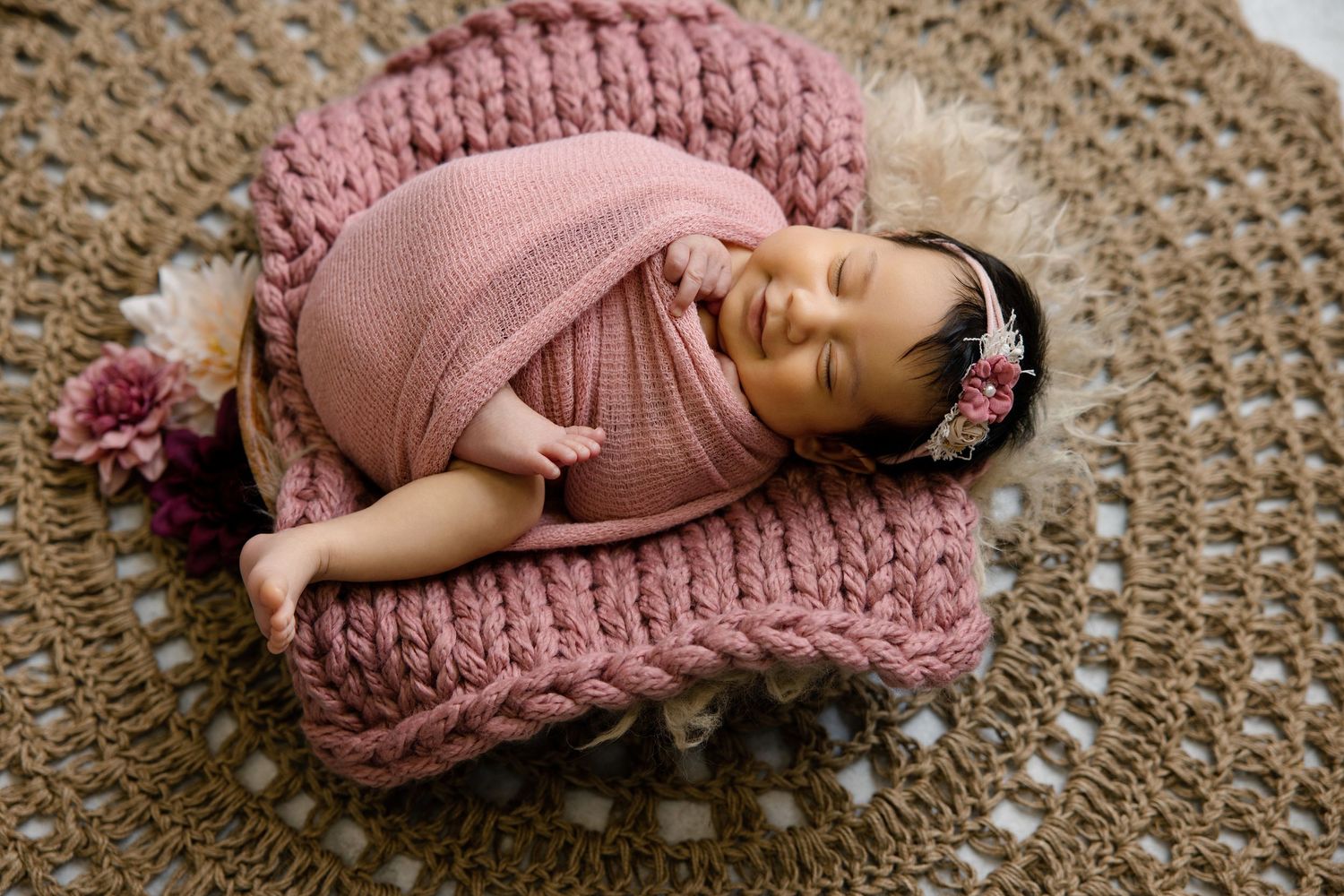 Newborn baby girl swaddled in pink knit wrap, sleeping peacefully in a cozy basket with floral headband.