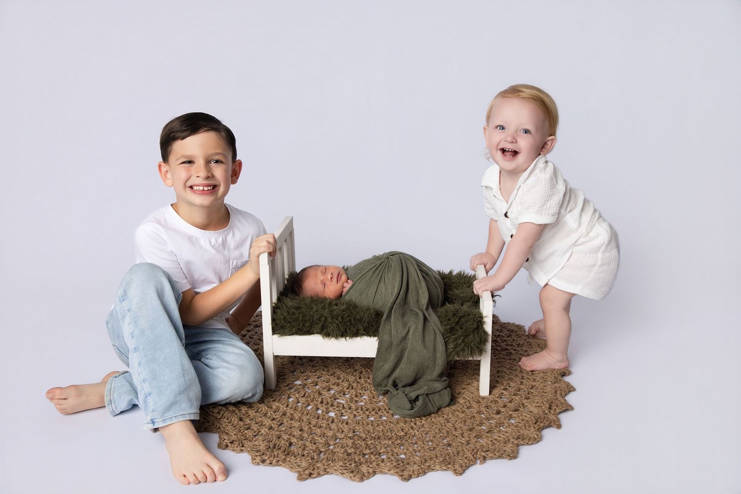 Two young siblings smiling beside their newborn baby wrapped in green on a tiny white bed.
