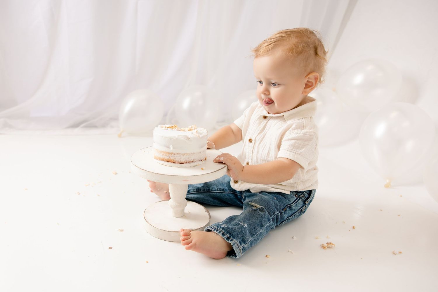 Baby boy in jeans and white shirt touches first birthday cake on white pedestal stand during cake smash photo session.