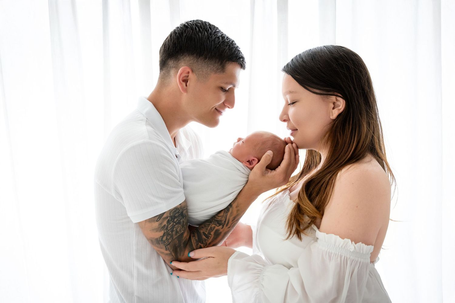 New parents tenderly holding their newborn baby together in a bright white studio setting.