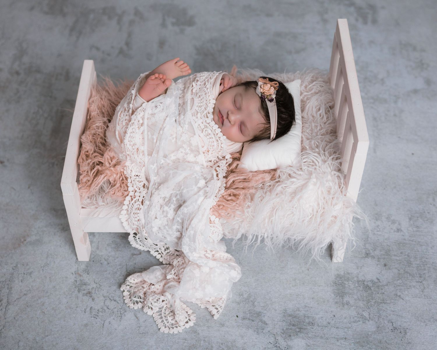 Newborn baby sleeping peacefully on a white miniature bed with lace wrap and floral headband in a professional photo.