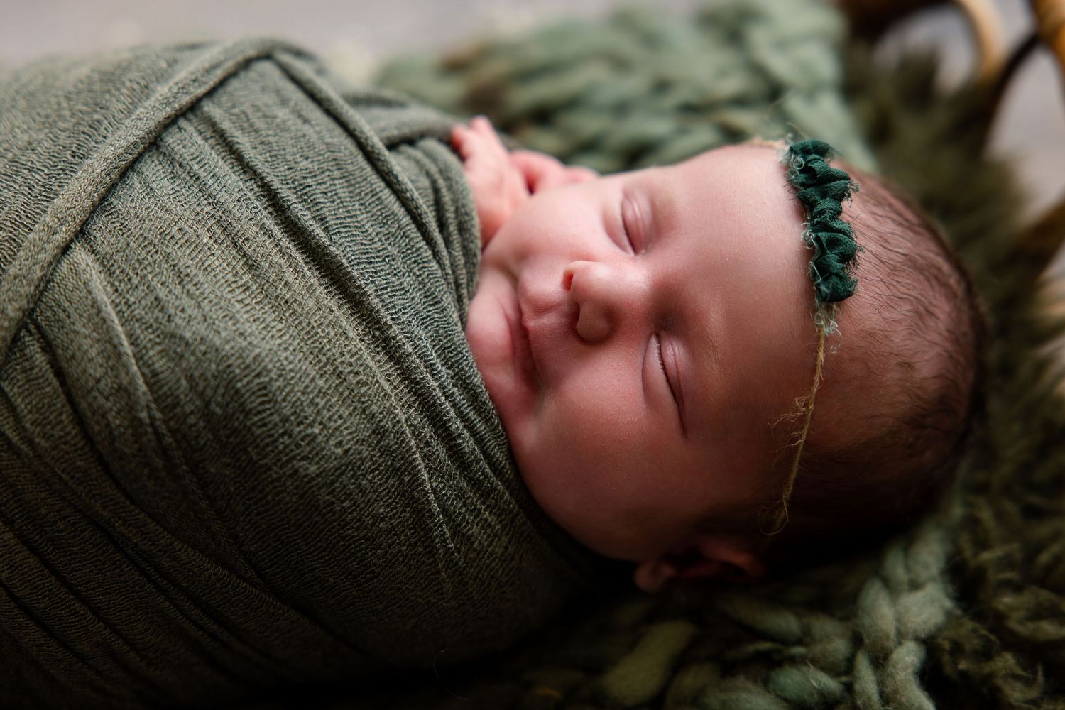 Sleeping newborn baby swaddled in green wrap with floral headband, peaceful infant portrait photography.