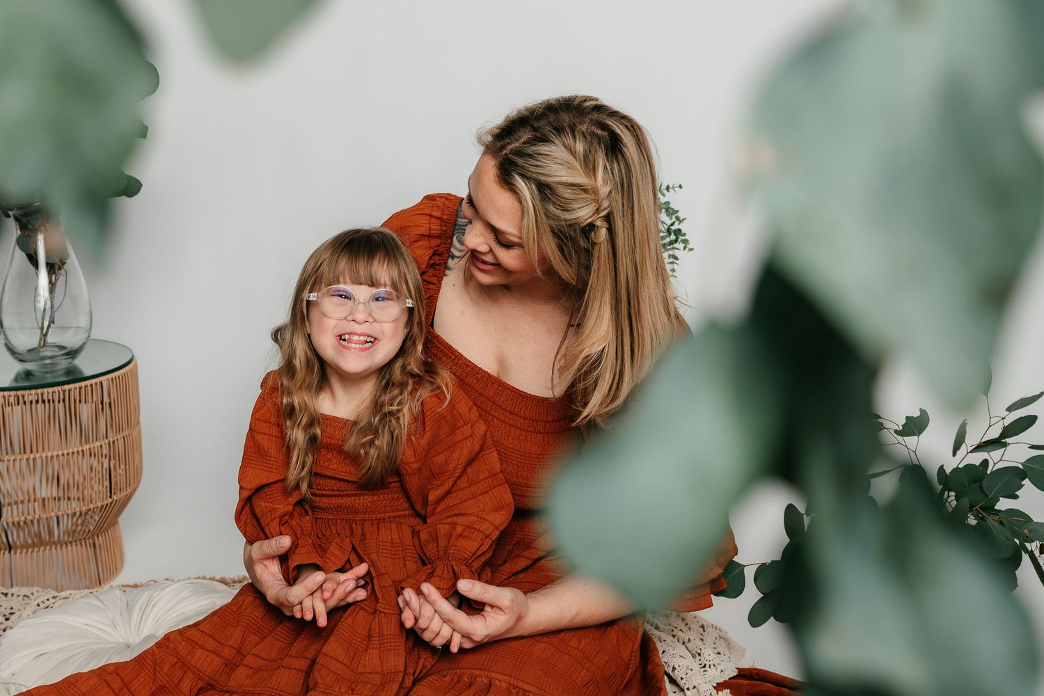 A mother and daughter in matching rust-colored dresses laugh together in a boho-styled indoor photo session in the Bonsall Studio
