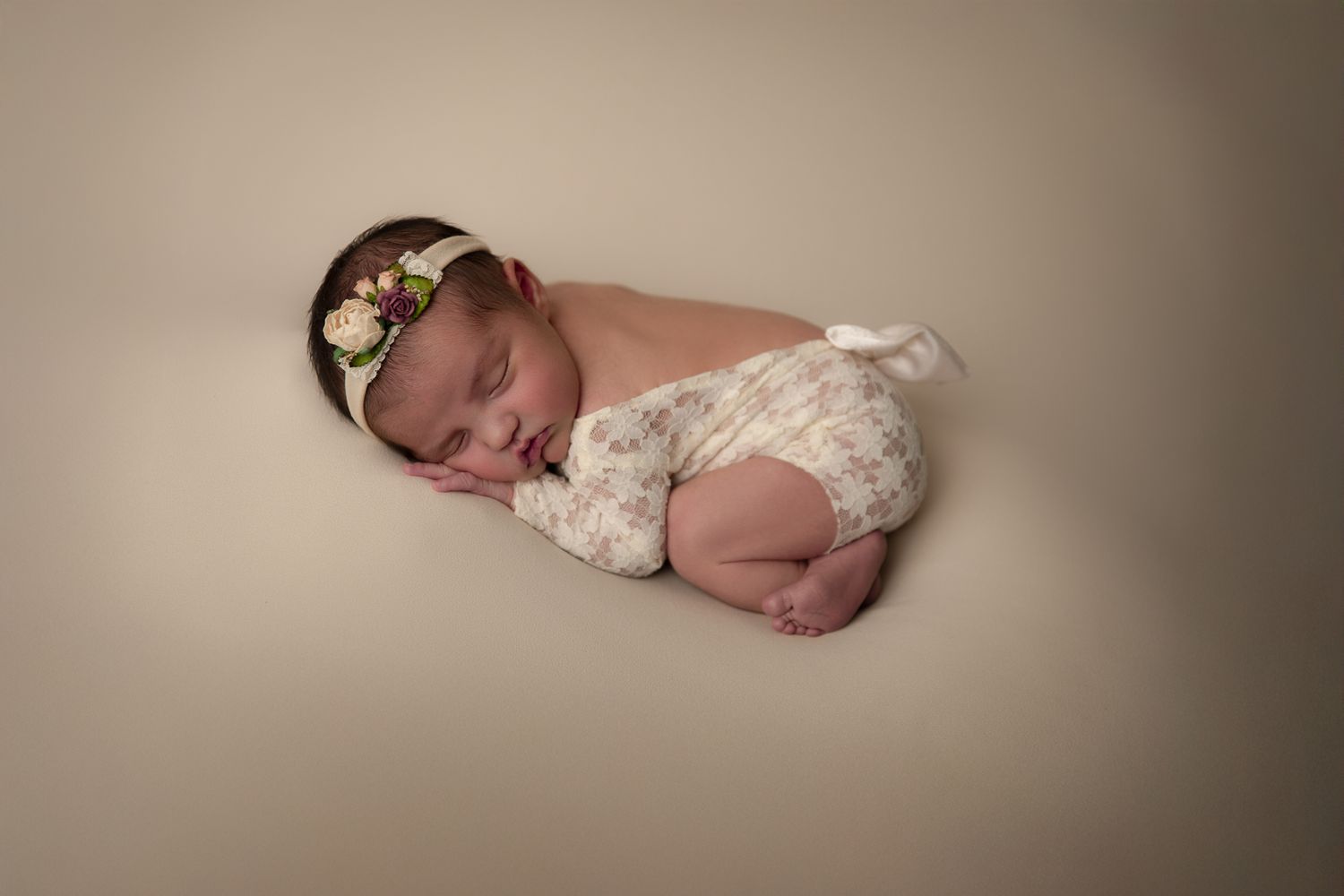 Peaceful newborn baby in floral romper with flower headband posed on beige backdrop. vista newborn photographer in Bonsall