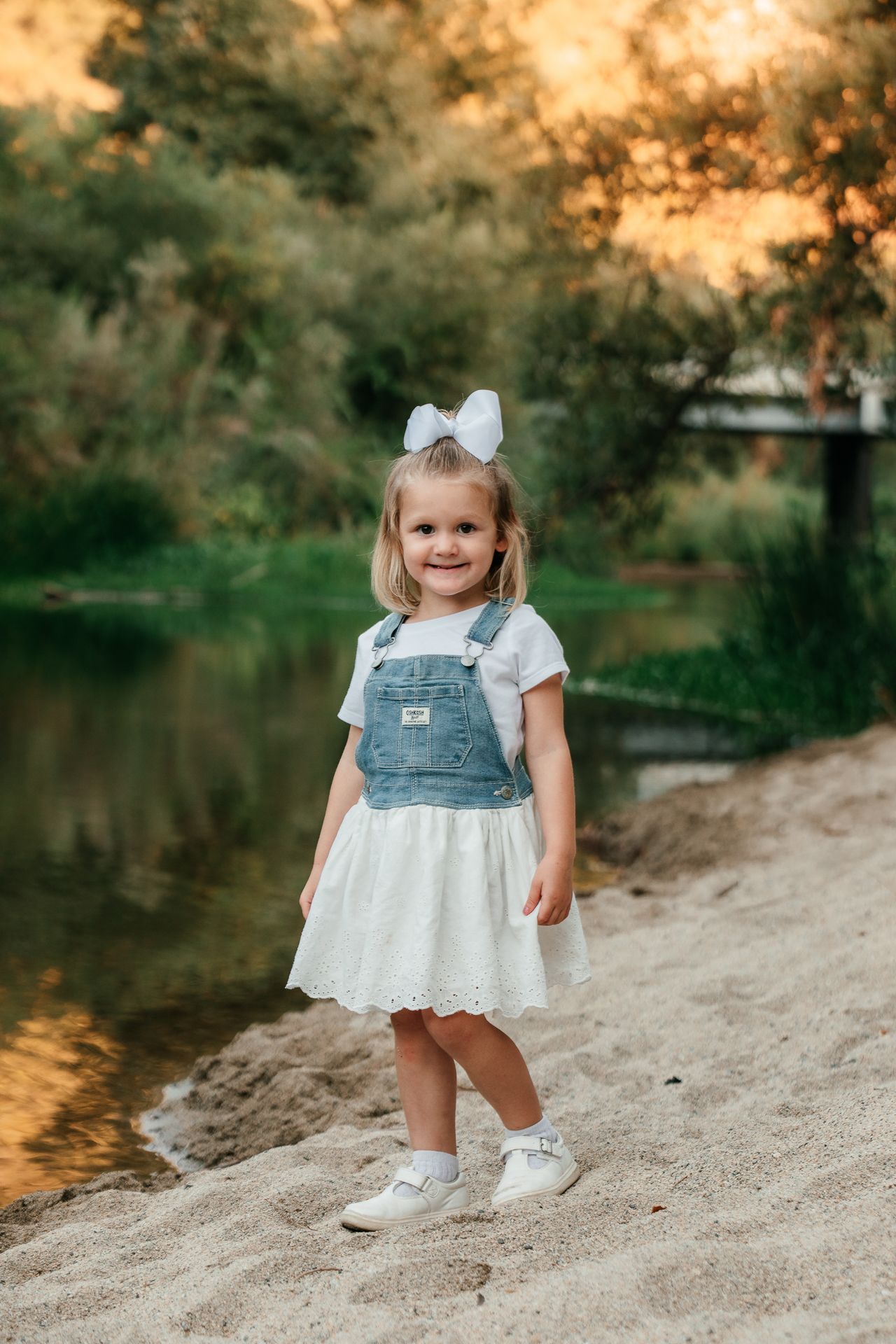 Adorable blonde toddler girl in denim overalls smiles on sandy riverbank at sunset.